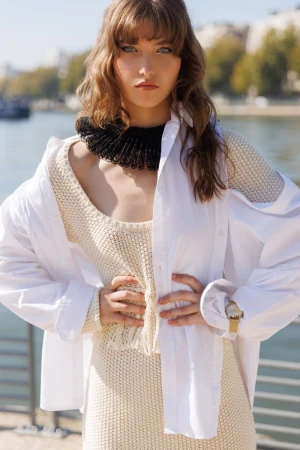 Outdoor fashion shot of a woman with wavy hair wearing a white shirt over a dress, with a beaded collar, posing near a river.