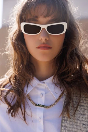 Close-up of a woman with wavy brown hair wearing sunglasses, a white shirt, and a gold necklace with the Lacoste logo.