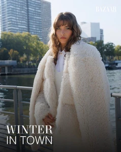 Model in white fuzzy coat standing by a riverbank with city buildings in the background for Harper's Bazaar Ecuador.
