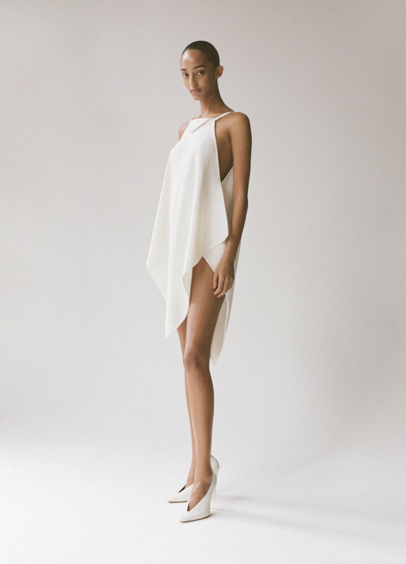 A slender female model poses in a draped white dress and white wedge heels against a neutral background, full body shot, fashion.