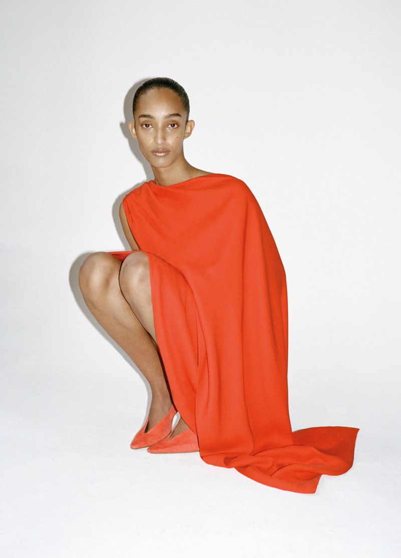A woman in a vibrant orange dress poses in a crouched position against a stark white backdrop, showcasing the dress's elegant design.
