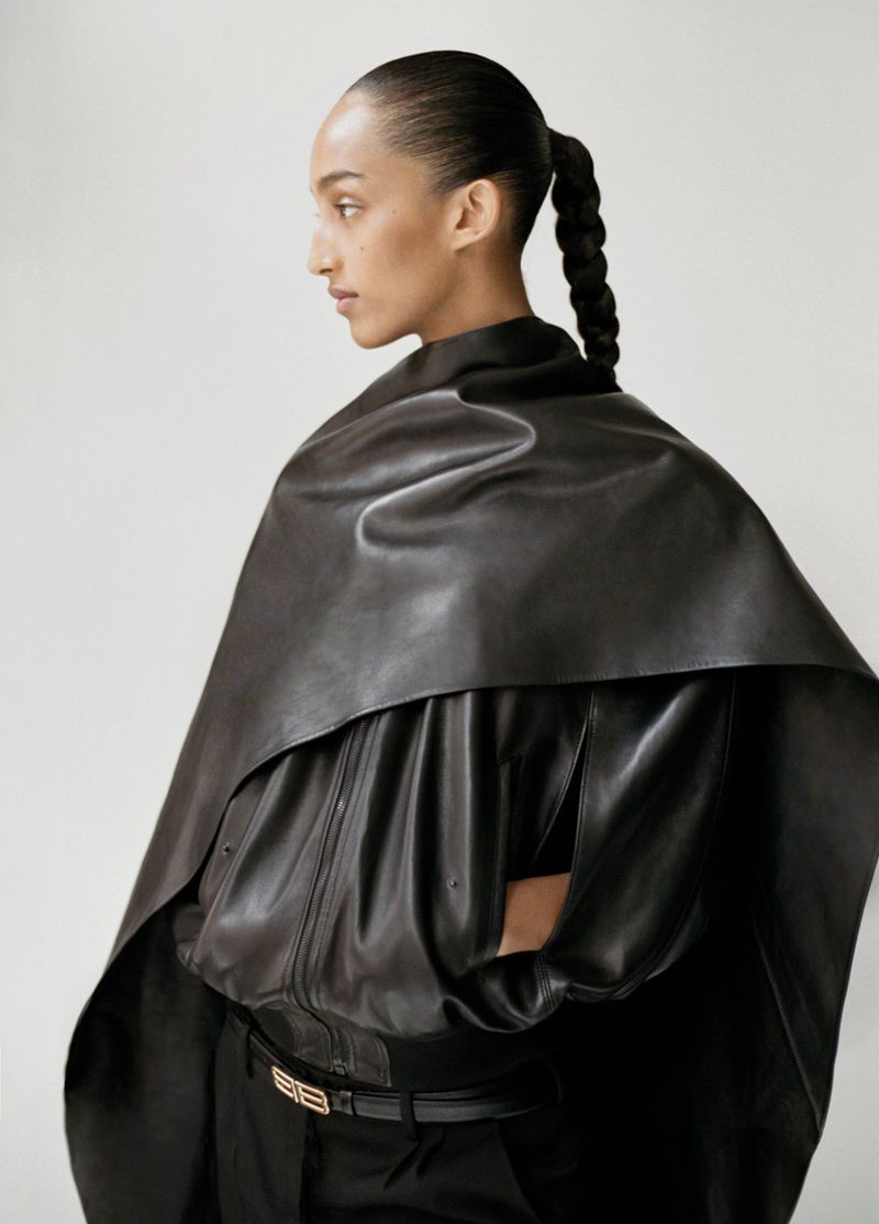 Profile shot of a woman wearing a black leather cape-like jacket and pants against a neutral background in a fashion-editorial setting.