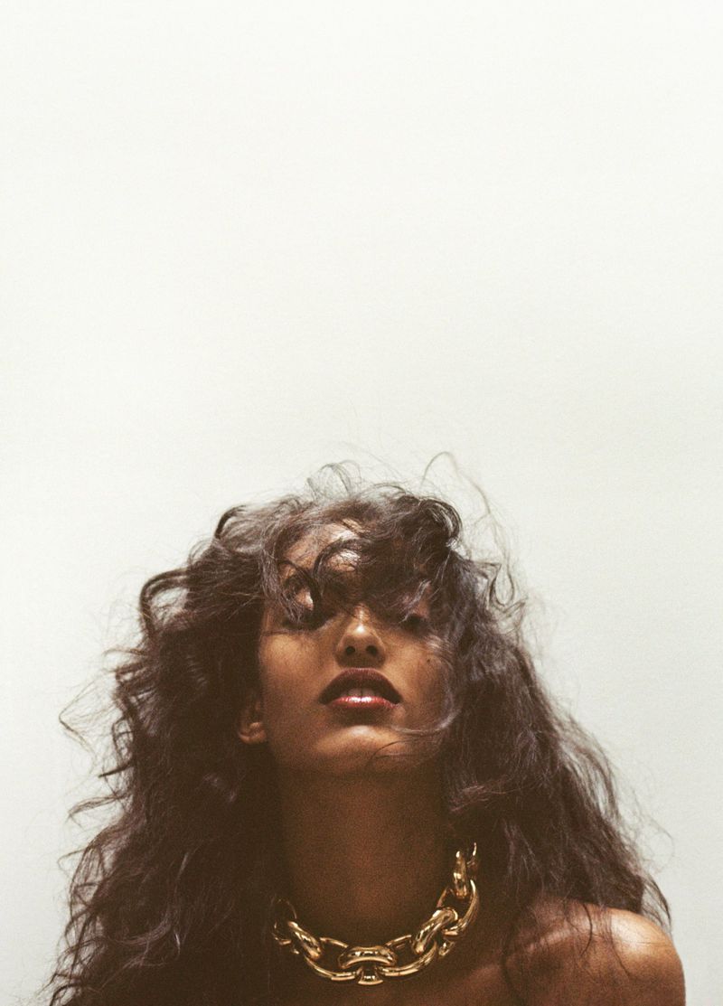 Portrait of a woman with curly dark hair and a gold chain necklace looking up against a white background in a studio setting.