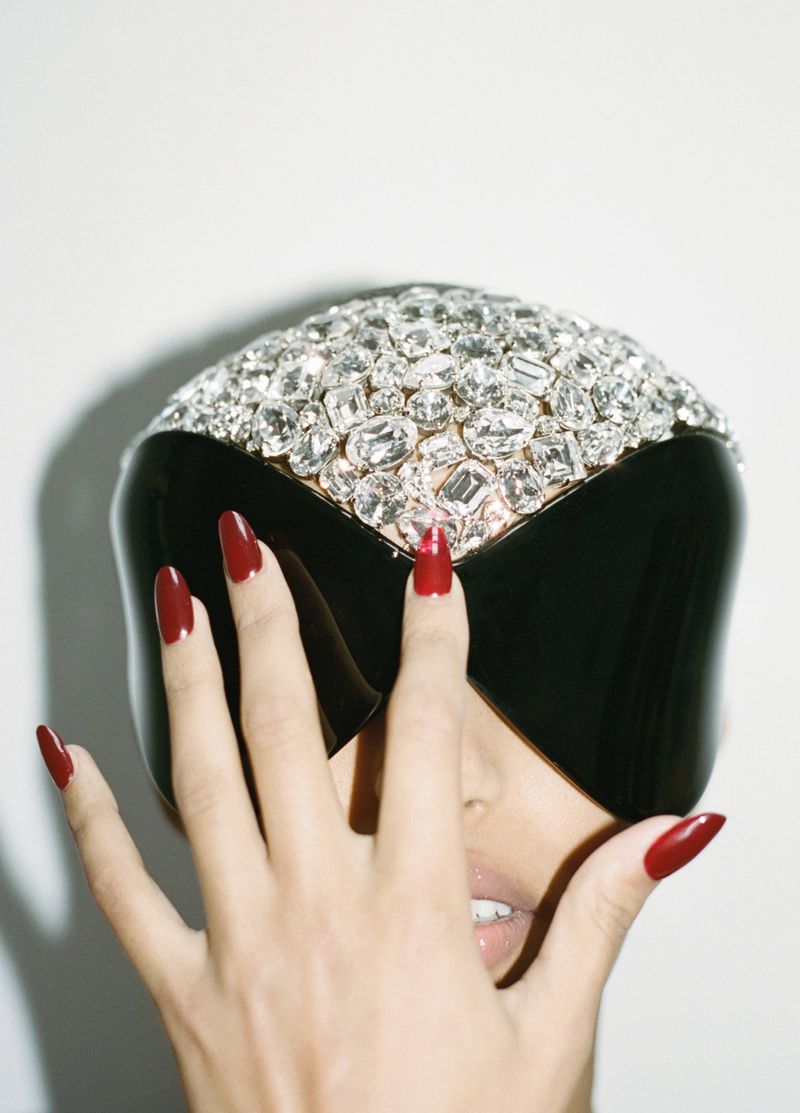 Woman with dark red manicure poses with a black visor covered in rhinestones in a studio setting against a plain white background.