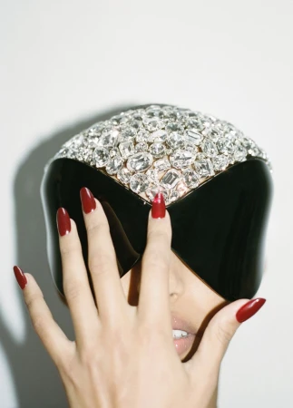 Woman with dark red manicure poses with a black visor covered in rhinestones in a studio setting against a plain white background.
