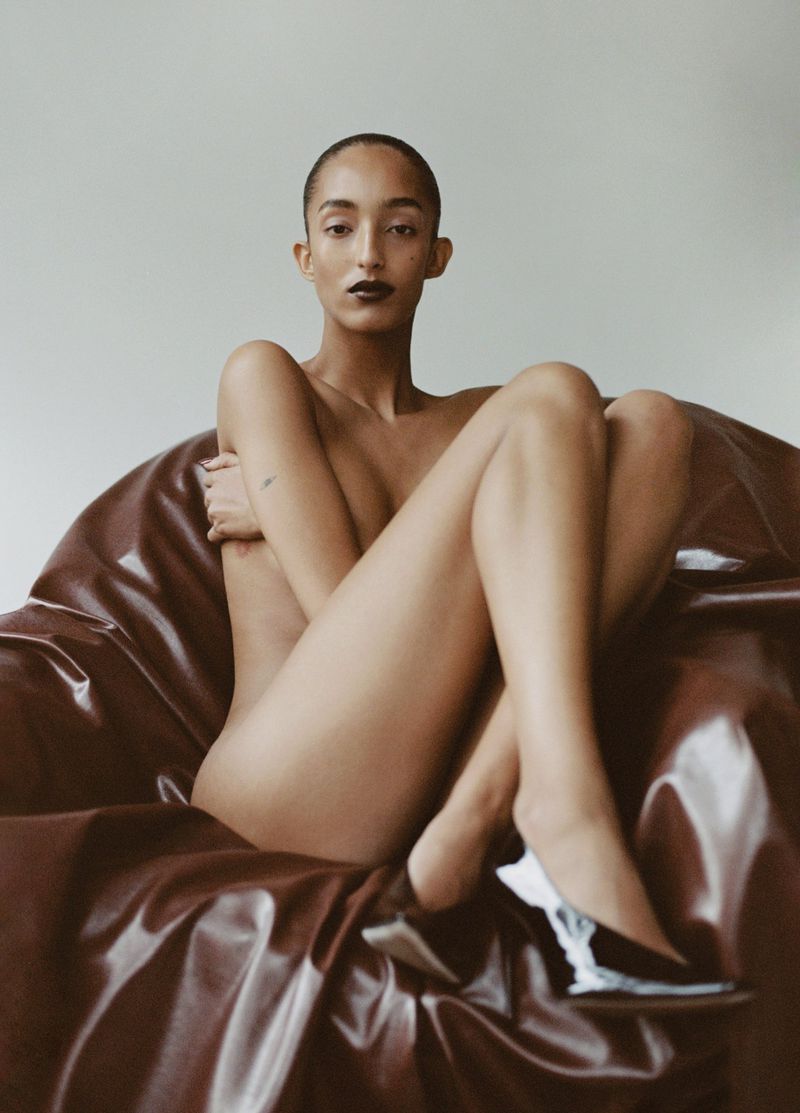 Woman with shaved head poses in studio sitting in a large brown leather chair, wearing black heeled shoes in beauty portrait.