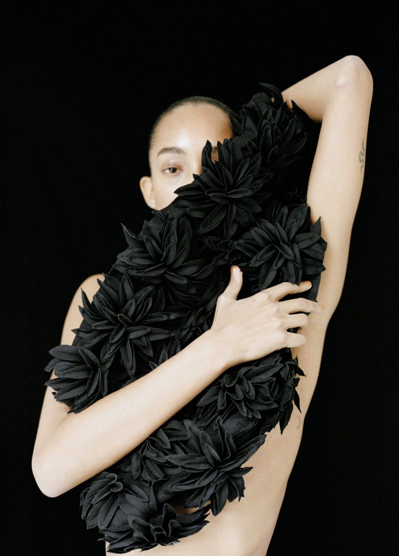 Woman embraces a black floral arrangement in a studio setting. Dark background emphasizes her natural features and dramatic floral piece.