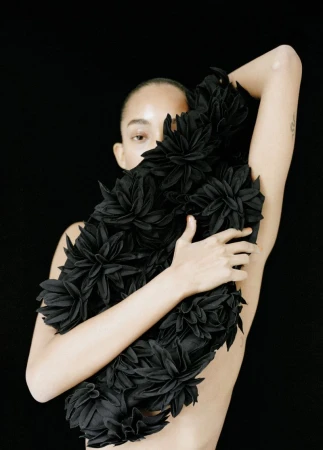 Woman embraces a black floral arrangement in a studio setting. Dark background emphasizes her natural features and dramatic floral piece.