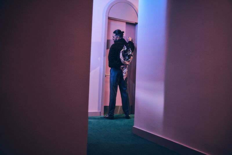 Adult male walks through doorway carrying wrapped flowers in interior hallway with pink walls and green carpet, back view.