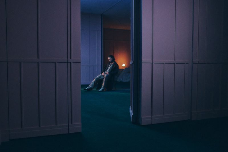 Adult male seated indoors on a teal surface, viewed through an open doorway, illuminated by an orange lamp, creating a contemplative mood.