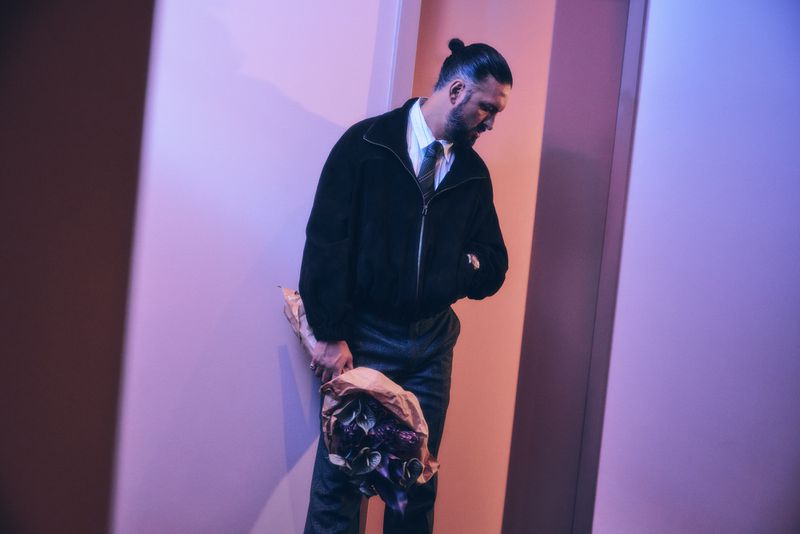 A man with a topknot hairstyle in a suit and black jacket holds a bouquet of flowers in a softly lit hallway or corridor.