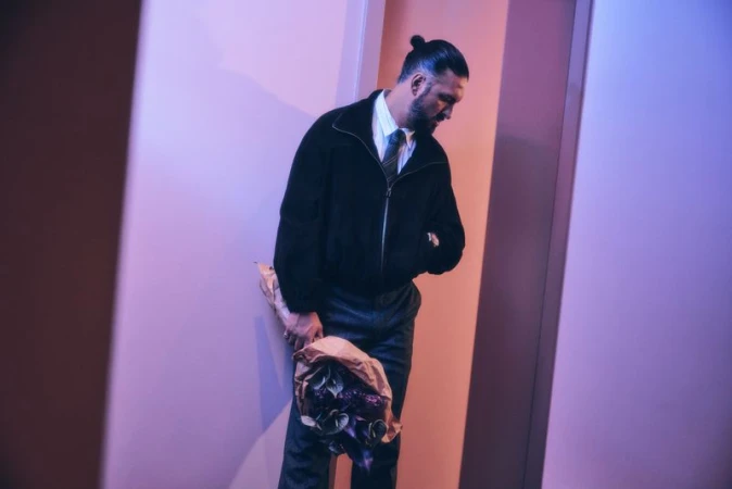 A man with a topknot hairstyle in a suit and black jacket holds a bouquet of flowers in a softly lit hallway or corridor.