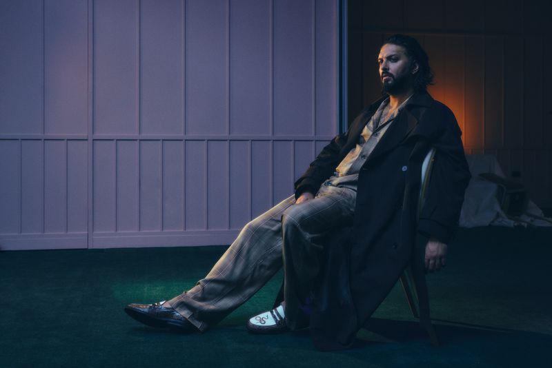 Adult man in a dark coat and patterned trousers seated on a chair in an indoor setting with violet walls and green carpet.