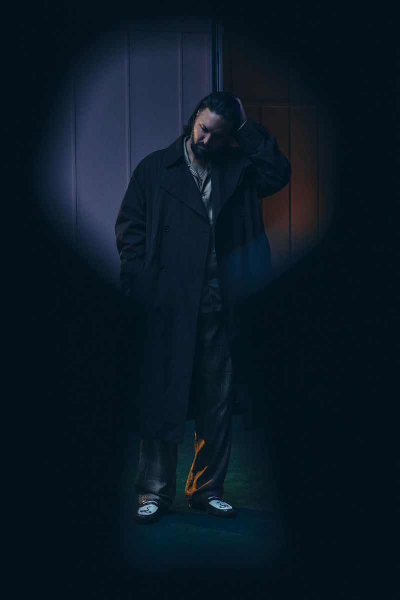 Portrait of a bearded man wearing a long dark coat and striped pants, standing indoors with a hand to his head.