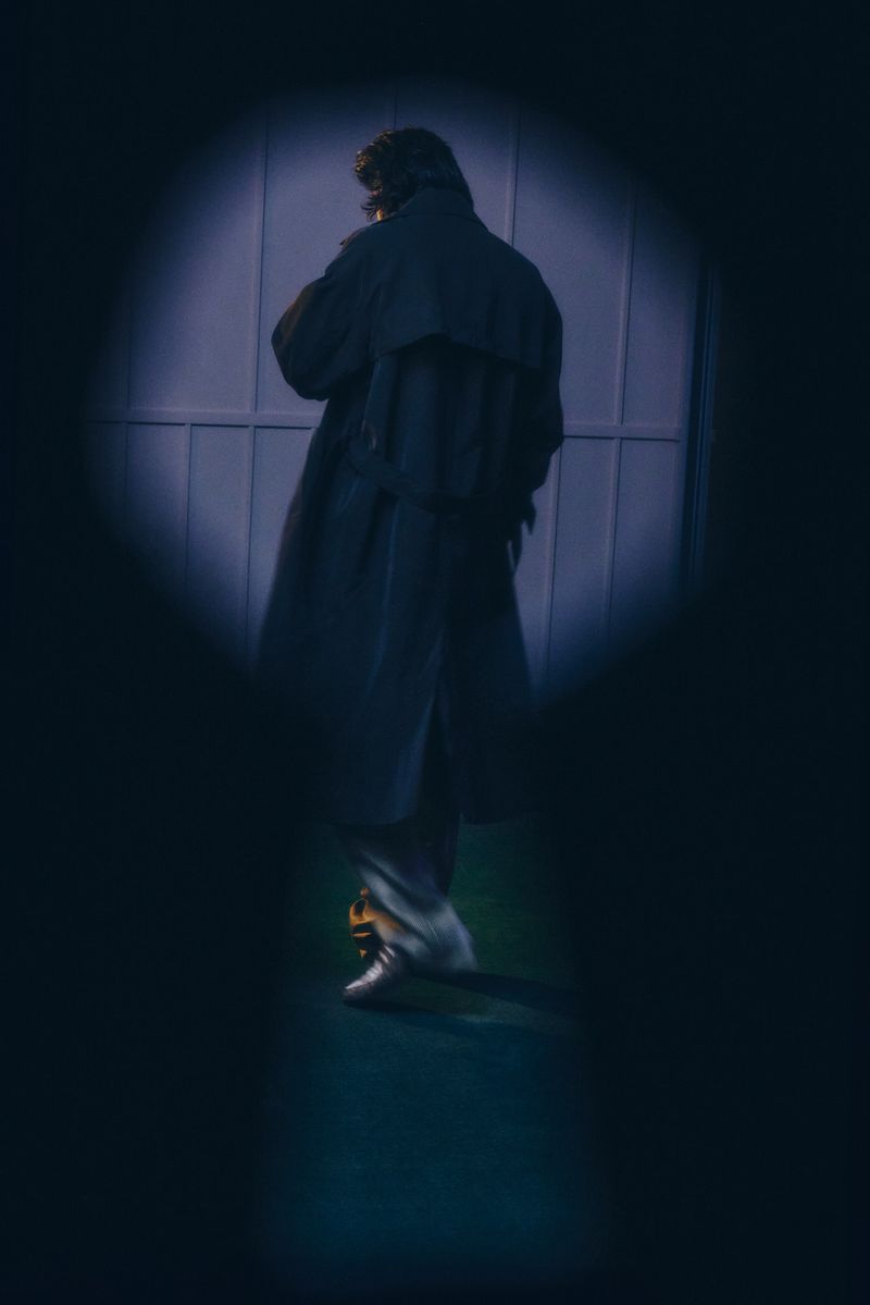 Man in a trench coat walks near a blue door, viewed through a dark circular opening, in dim, moody lighting.