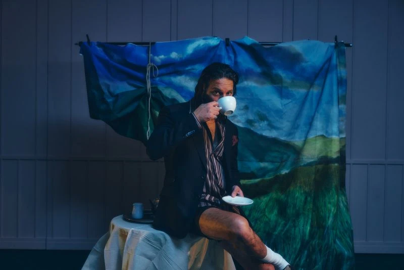Man in blazer and pajamas drinks tea, seated before a mountain landscape painting in a dimly lit indoor setting with white wainscoting.