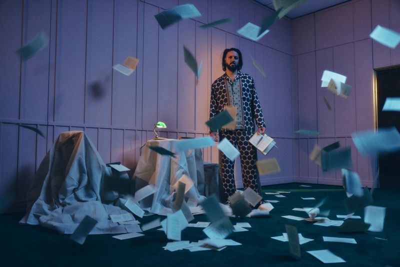 Man in patterned pajamas surrounded by floating papers in a dimly lit room with vintage decor and a small green lamp on a covered table.
