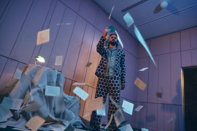 Man in polka-dot pajamas stands surrounded by floating book pages inside a room with a lamp on a draped table.