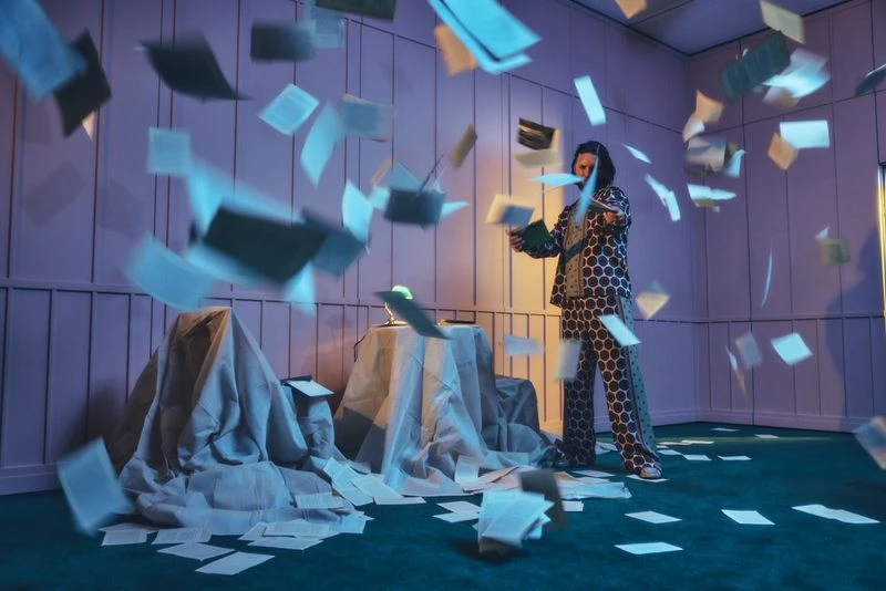 Adult male in patterned pajamas stands amid a flurry of papers in a room with green carpet and lavender walls, creating an organized mess.