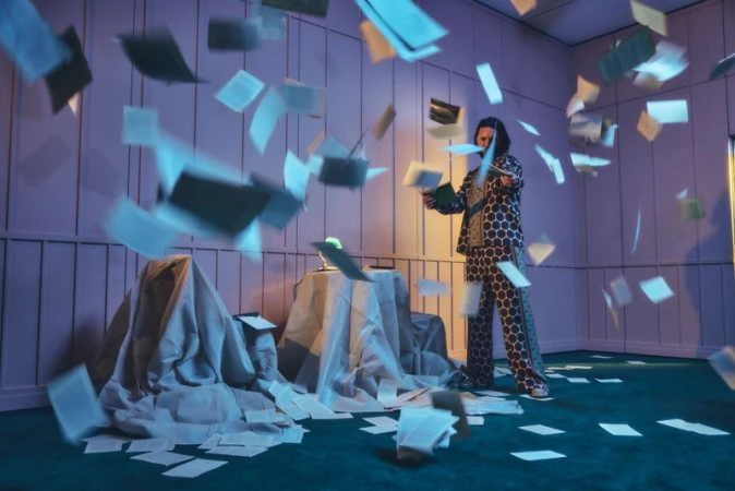 Adult male in patterned pajamas stands amid a flurry of papers in a room with green carpet and lavender walls, creating an organized mess.
