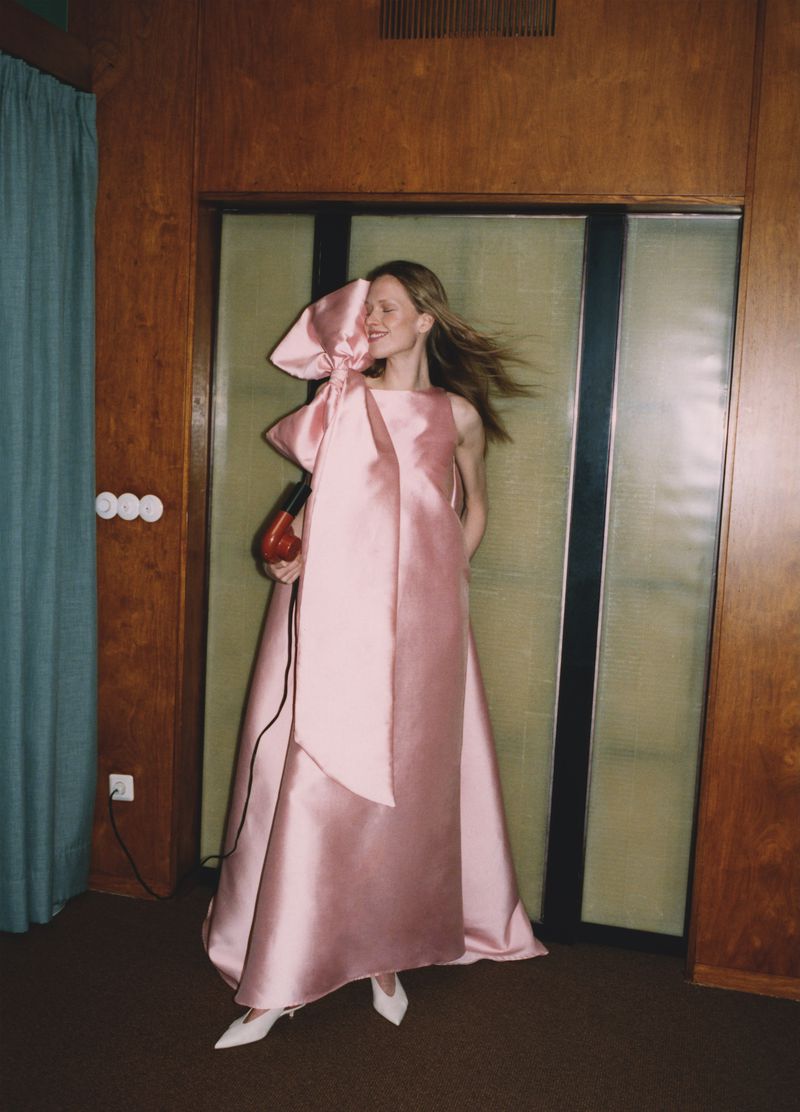 Woman in a pink gown with a large bow holds a red hair dryer in an indoor setting with wood-panelled walls and white heels.