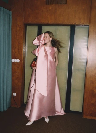 Woman in a pink gown with a large bow holds a red hair dryer in an indoor setting with wood-panelled walls and white heels.