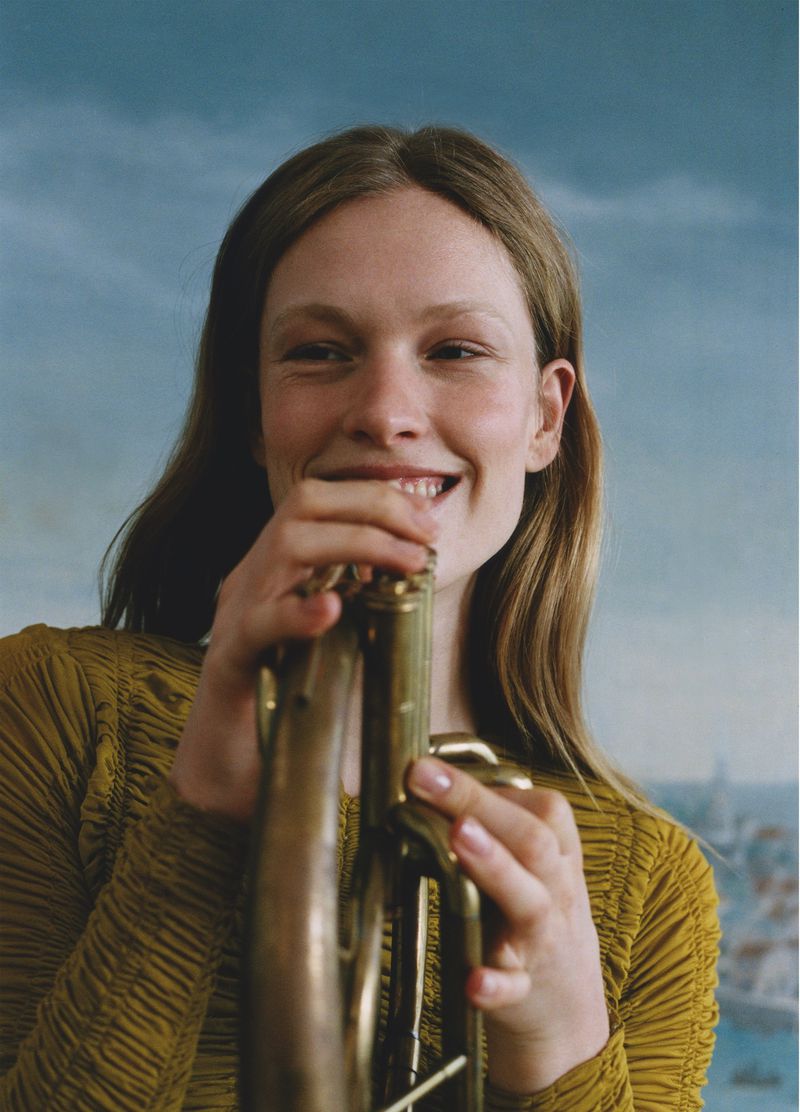 Young woman smiling while holding a brass trumpet in front of a sky-painted background, creating a cheerful and classic portrait.