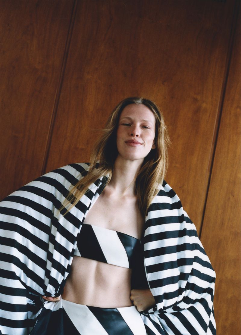 A fair-skinned woman with long blonde hair wearing a black and white striped outfit poses indoors against a wood paneled wall.