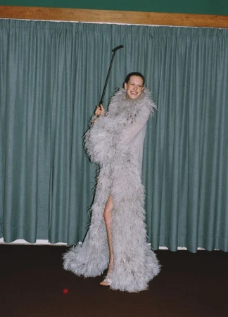 A woman in a feathered gown poses indoors, holding a golf club, with a red golf ball on a dark brown floor and a blue curtain background.