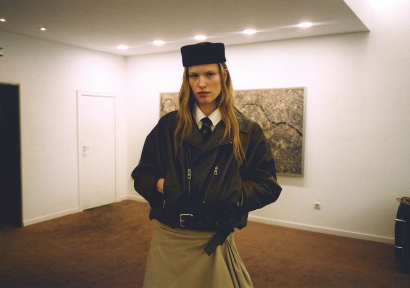 Adult male fashion model with long hair poses in a bomber jacket, tie, kilt, and black cap in a minimalist indoor setting.