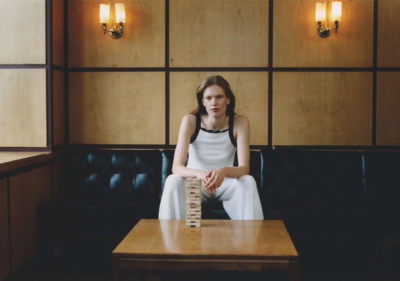 Woman sits near Jenga tower in a room with wood paneled walls, black leather couch, and warm lighting from wall sconces.