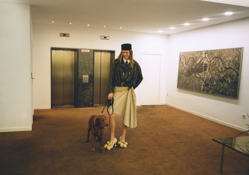 Woman in olive jacket and hat walks a brown dog on a leash, standing on a brown carpet, with an elevator bank in the background.