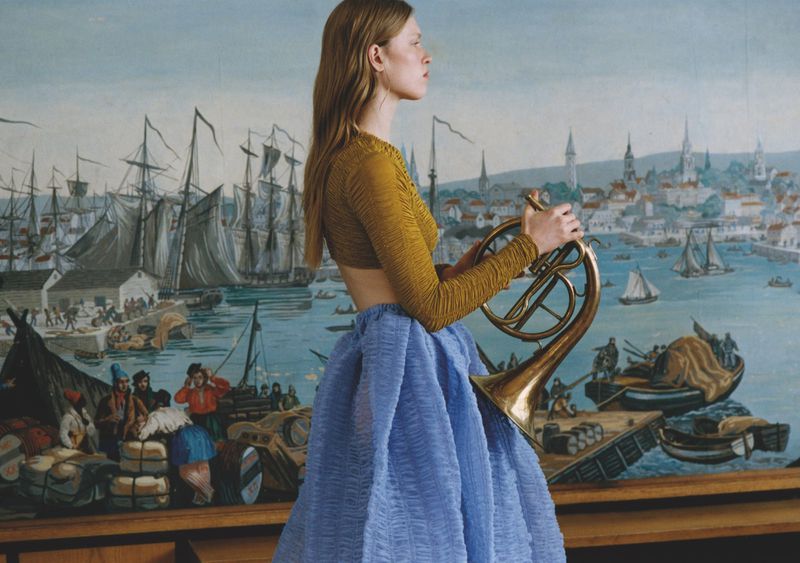 Fashion shot of a woman holding a brass French horn, wearing a light blue ball skirt and a mustard-colored crop top against a mural backdrop.