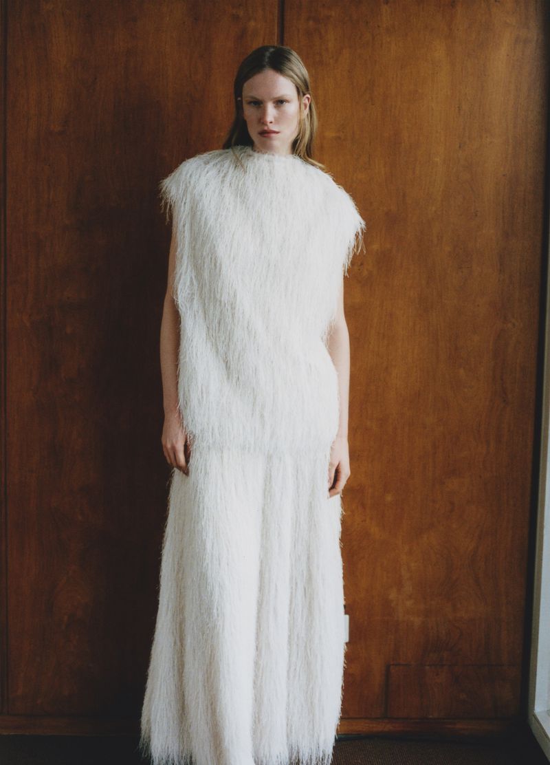 Fashion shot of a woman in a white, fringed, sleeveless top and wide-legged trousers posing against a brown wood-paneled wall.