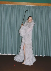 Model wearing a feathered gray coat holding a golf club standing in front of green curtains for Vogue Turkey