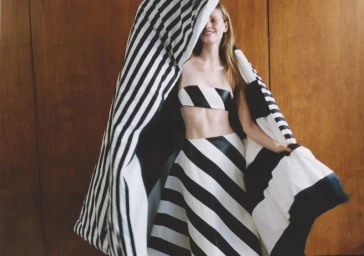 Model wearing a black and white striped strapless top and skirt holding up a matching striped fabric against wood paneling