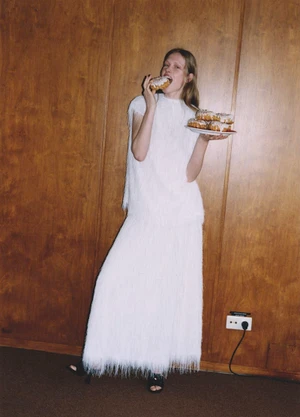 Model in white fringed dress eating a frosted pastry while holding a plate of donuts against wood paneling
