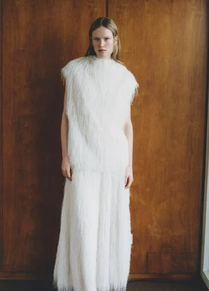 Model Leanne De Haan wearing a white fringed textured dress standing against a dark wood paneled wall for Vogue Turkey.