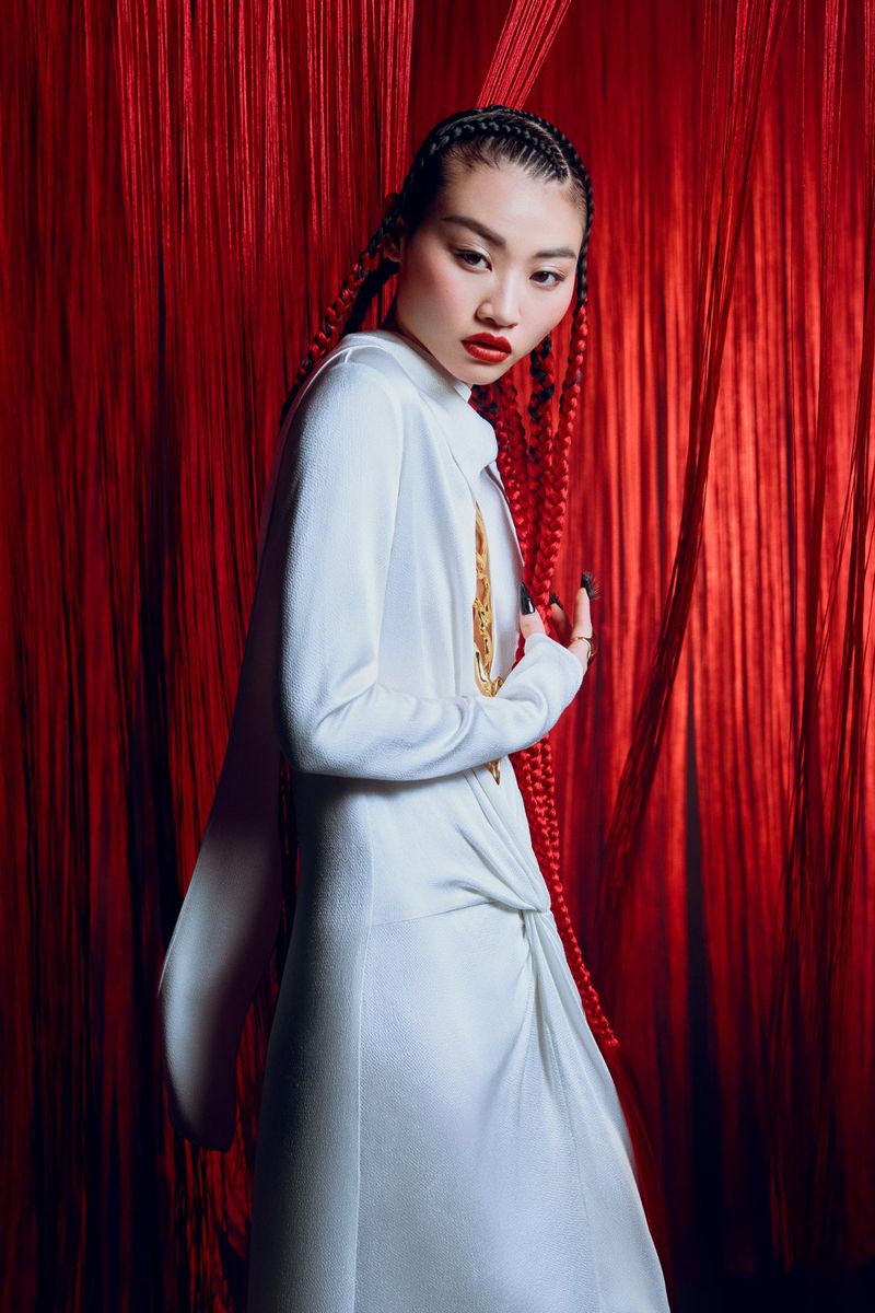 Fashionable Asian woman with red braided hair poses in a white dress against a red curtain, showcasing editorial style and studio lighting.