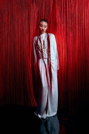 An Asian female model in white dress poses dramatically amidst a red rope curtain and a dark, glossy floor, showcasing fashion and style.