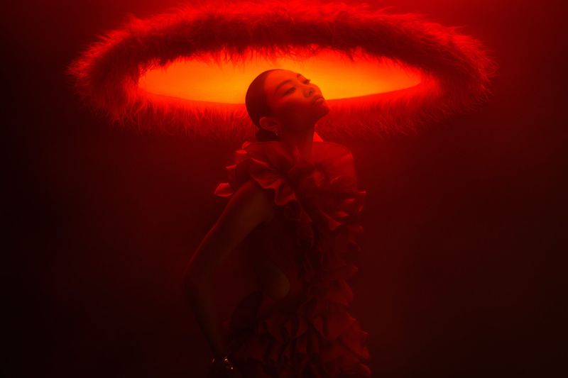 Adult female with dramatic make-up and ruffled garment in a red-lit room with soft overhead light, eyes closed, looking peaceful.