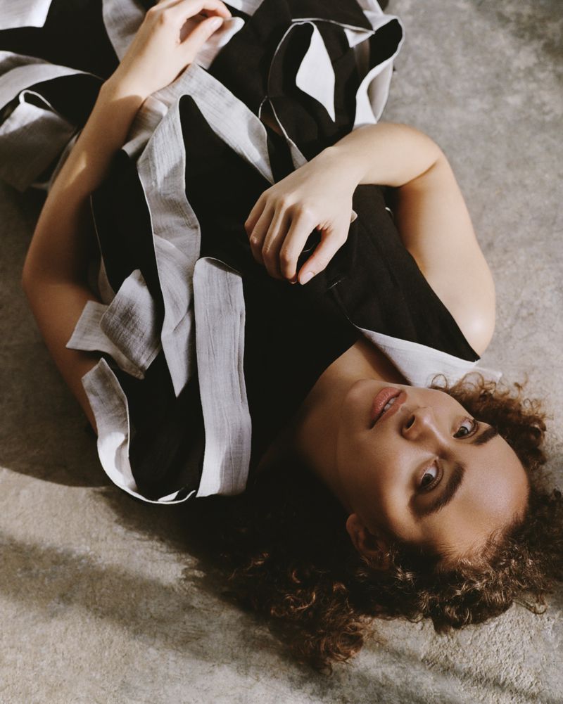 Overhead shot of woman lying on a textured surface wearing a flowing black and white striped dress, looking towards the camera.