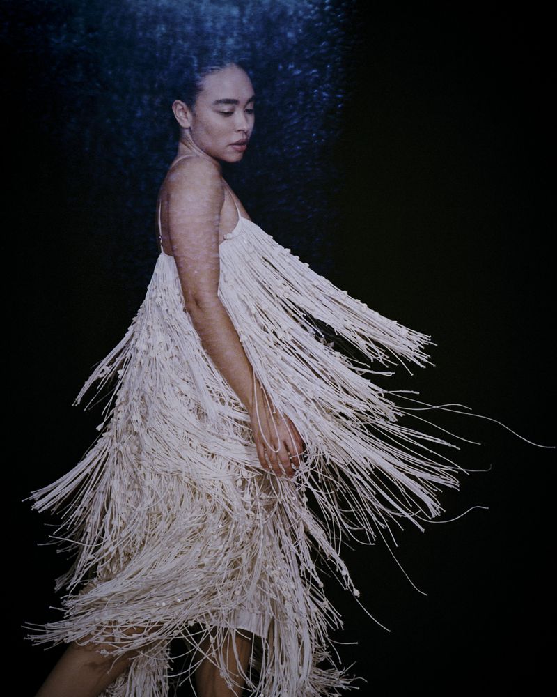 Woman in a cream-colored fringe dress, shot in studio with a black backdrop. The light catches the fringe details and highlights the model's skin.