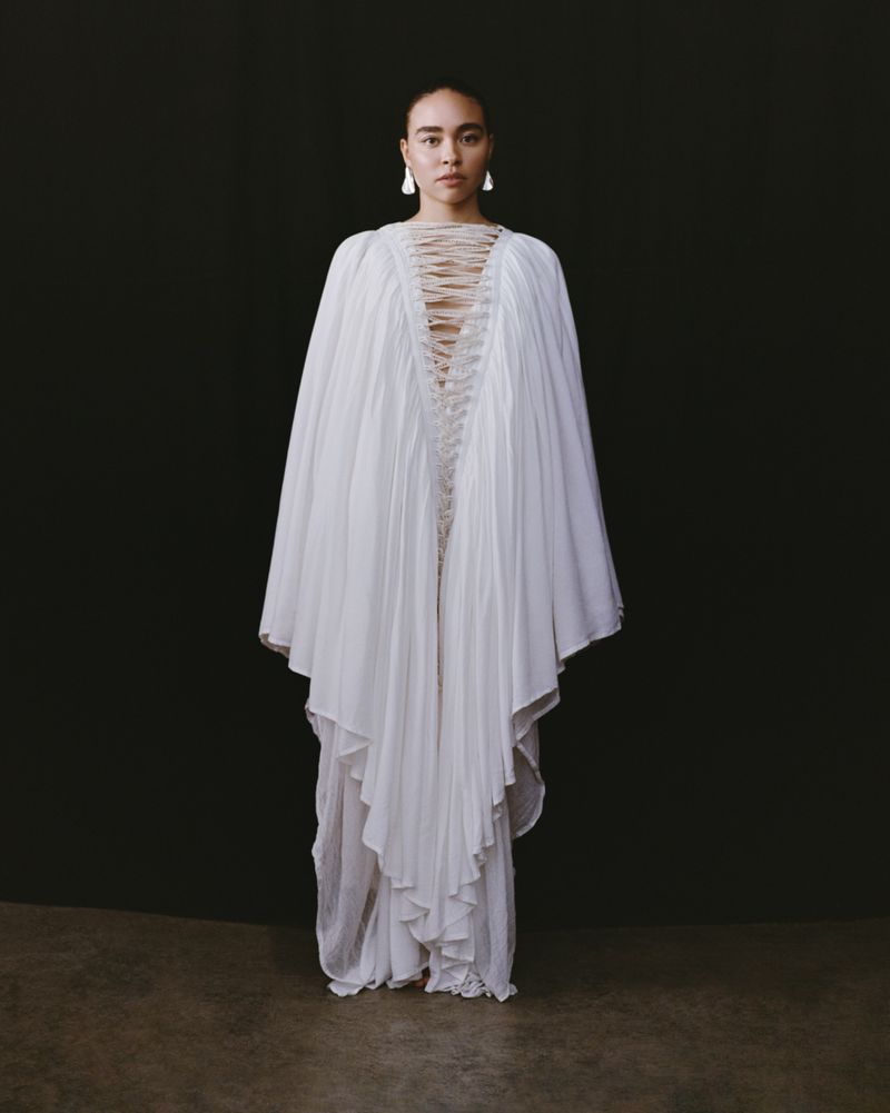 Woman in a flowing white gown with cape-like sleeves stands against a dark backdrop, earrings visible, shot in a studio setting.