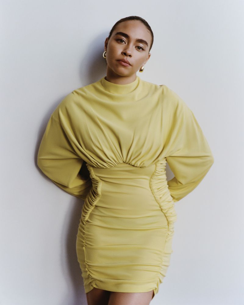 Woman in a butter-yellow mini dress poses in front of a white wall, wearing gold hoop earrings, in a studio shot for fashion.