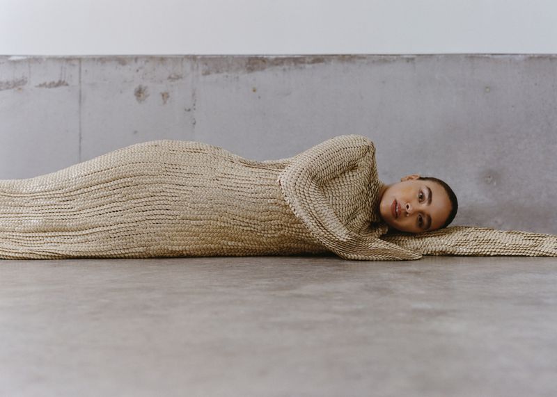 A woman lies on a gray surface wearing a textured beige dress. The shot is a medium shot with a concrete background.