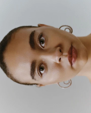 Close-up portrait of a woman with short hair and earrings against a neutral background, captured with soft lighting.