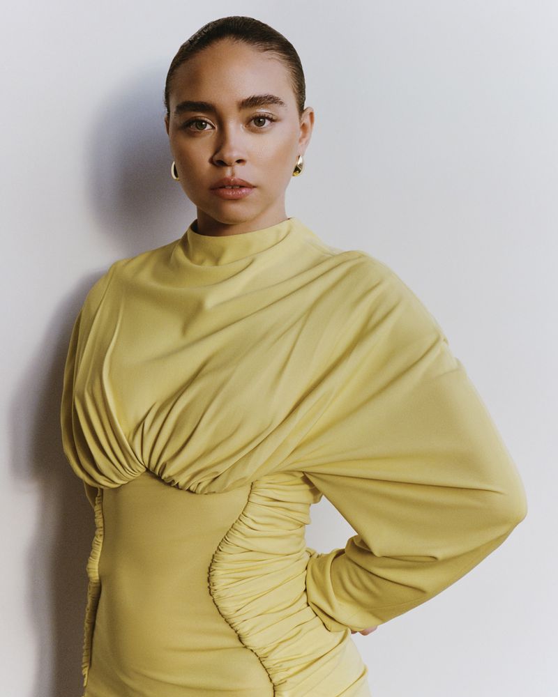 Portrait of a stylish young woman with dark hair wearing a fashionable yellow dress in front of a plain white background.