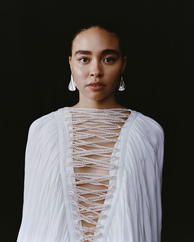 Portrait of a woman with fair skin and brown hair, wearing teardrop earrings and a white dress with lace details on a dark background.
