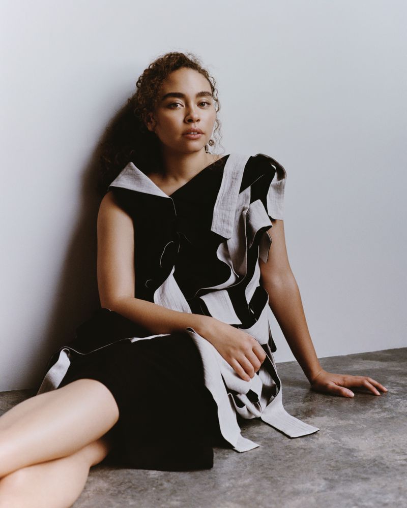 Portrait of an adult female model in a black-and-white dress with ruffled detail, seated in a photo studio with grey background.